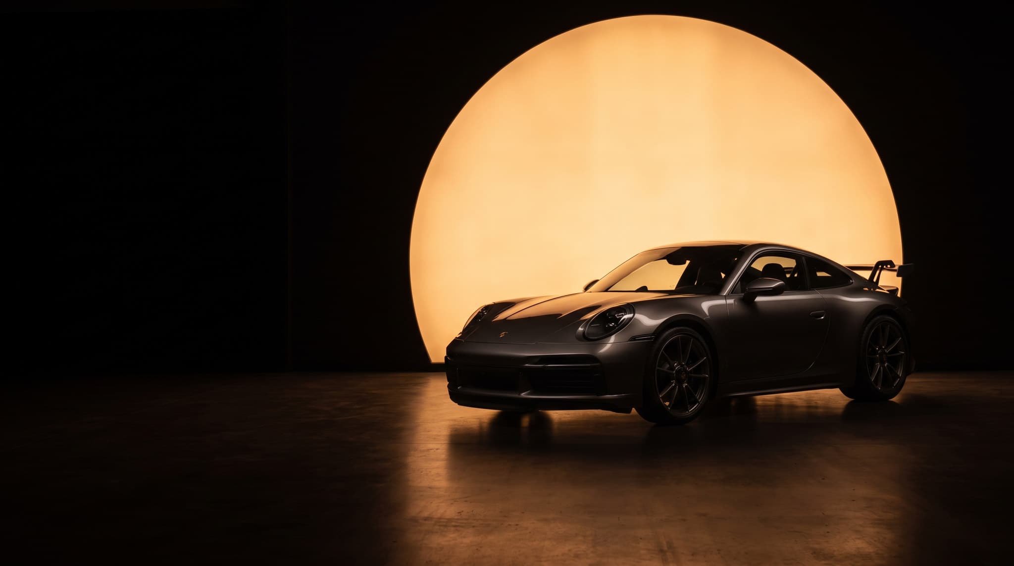 A single performance car in a darkened showroom at night, lit from behind by one large disc of warm amber light on the rear wall